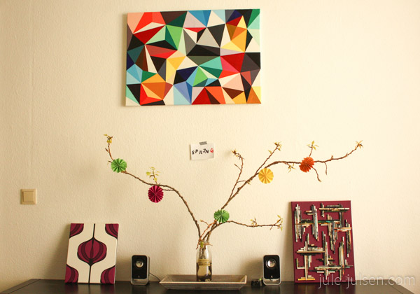 Sideboard mit selbst gemalten Bildern und einem Strauß aus Zweigen an dem bunte Papieronamente angehängt sind Sideboard mit selbst gemalten Bildern und einem Strauß aus Zweigen an dem bunte Papieronamente angehängt sind